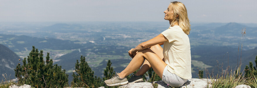Frau sitzt auf einem Stein in der Sonne, im Hintergrund Landschaftspanorama.