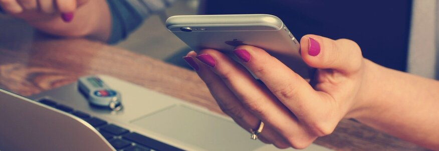 Eine Dame sitzt vor Ihrem Laptop mit dem Mobiltelefon in der linken Hand.